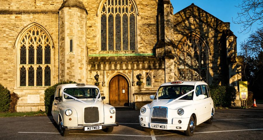 Pair of white wedding taxis with ivory ribbons outside a church